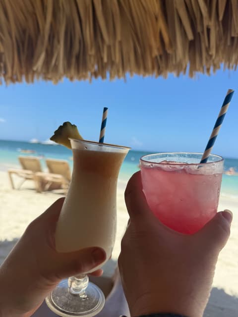 Two tropical cocktails raised in a toast under a thatched umbrella on a beach in Jamaica