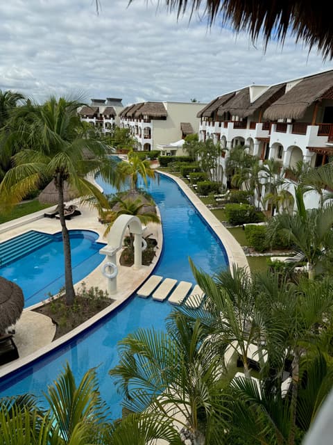 A winding pool lined with palm trees and thatched-roof suites at Valentin Imperial Riviera Maya
