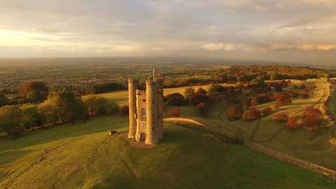 Broadway Tower on its grassy hilltop in golden evening light, with autumn trees and the Cotswold countryside stretching to the horizon