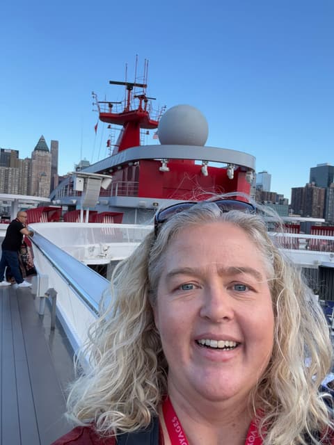 Michelle smiling on the top deck of a Virgin Voyages ship, with its red funnel and the New York City skyline behind her