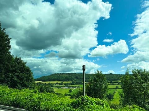 Green Cotswold farmland and wooded hills under a bright blue sky with big white clouds