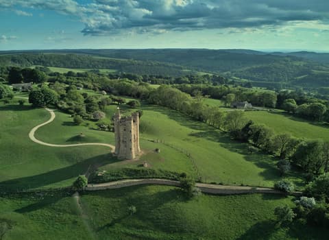 Aerial view of Broadway Tower on a grassy hilltop, with green Cotswold countryside rolling away behind it